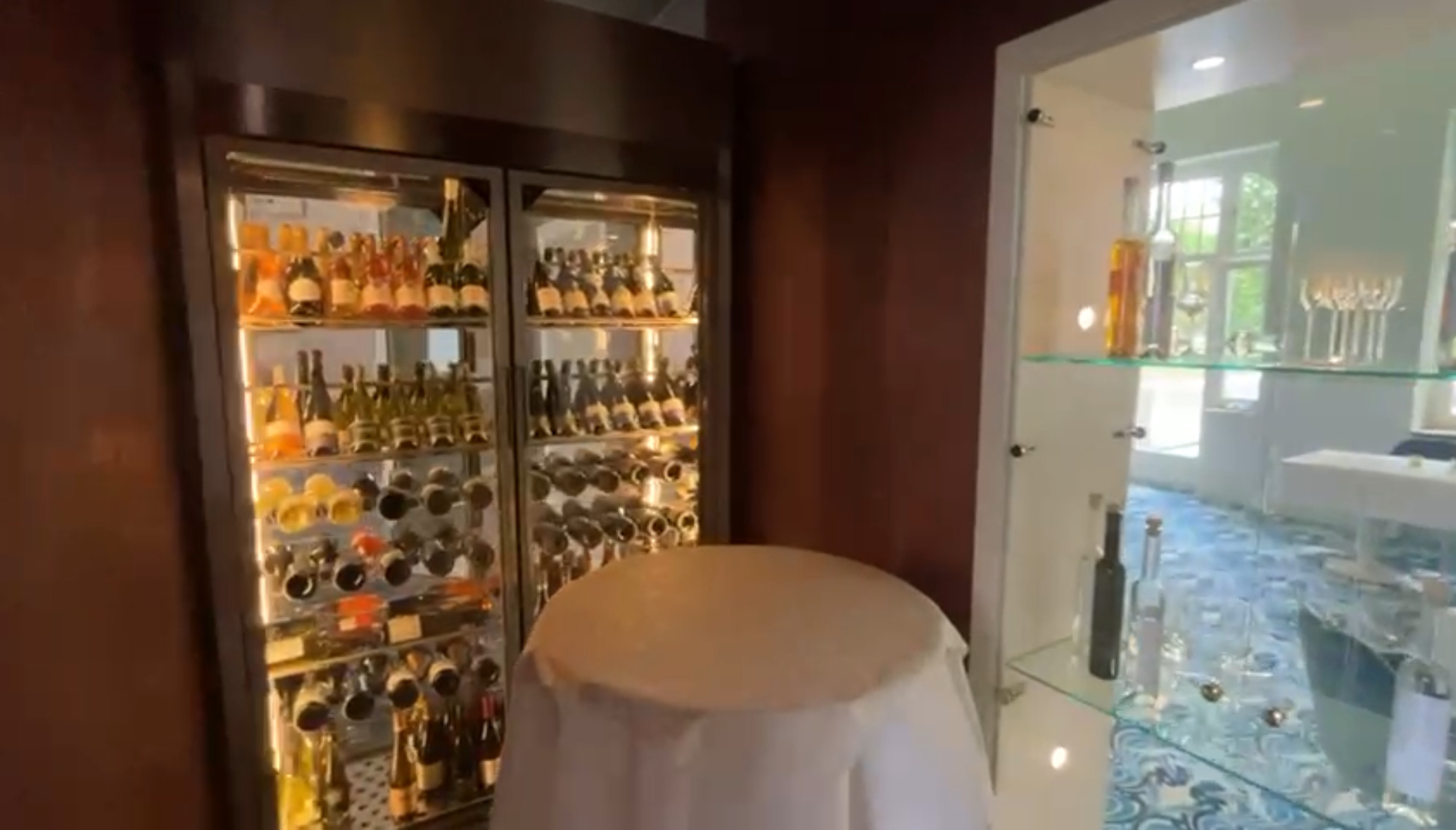 Ein gekühlter Weinschrank mit Glastüre, gefüllt mit Weinflaschen. Ein weißer Stehtisch und eine Vitrine aus Glas in der Wand eingelassen.